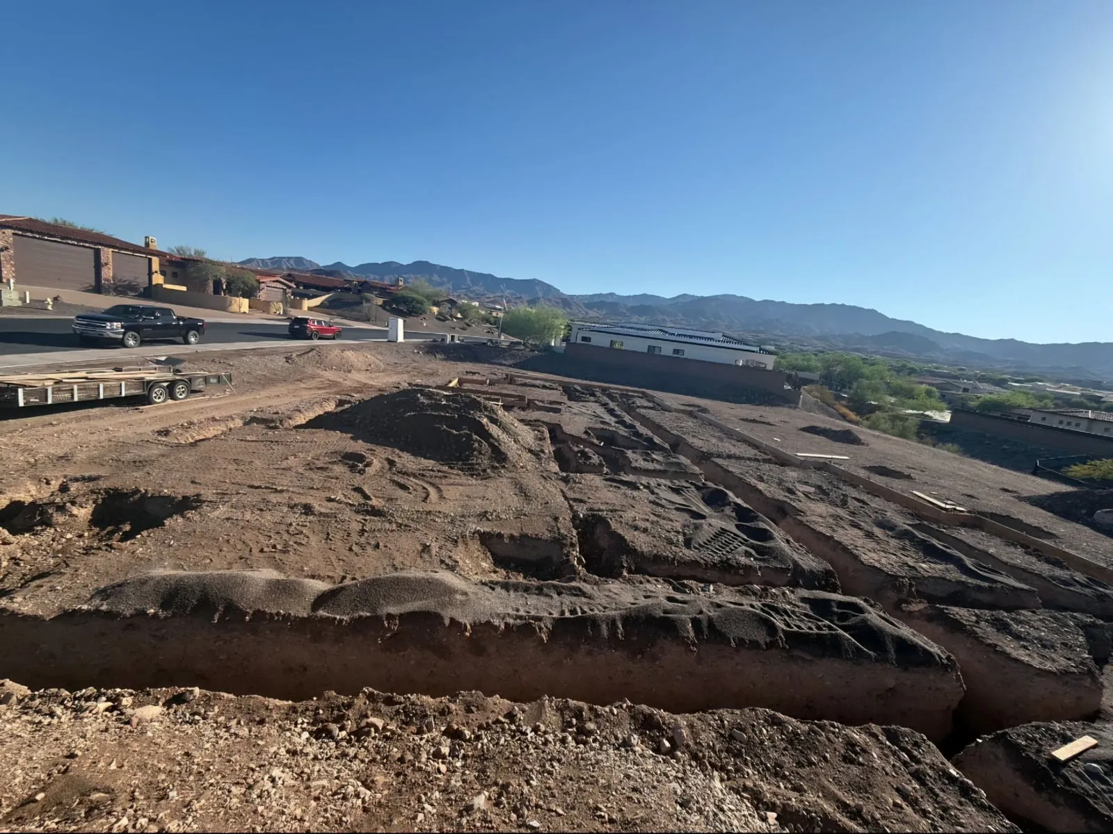 A new home site with open plumbing trenches, with desert homes and mountains behind
